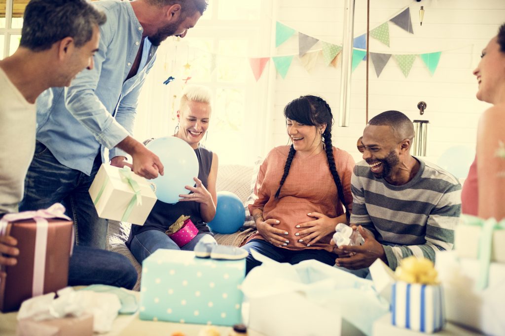 A baby shower with a bunch of people laughing and presenting gifts to a pregnant woman. 