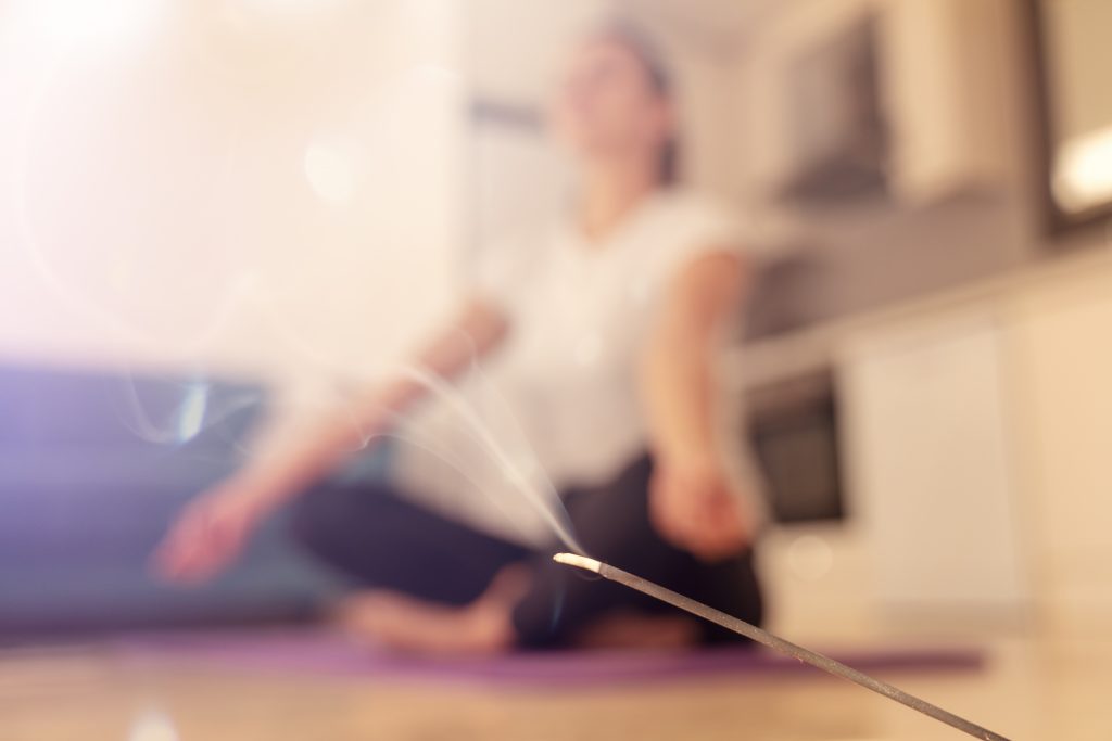 A burning incense stick pictured against  a blurry background of a woman doing yoga at home. 