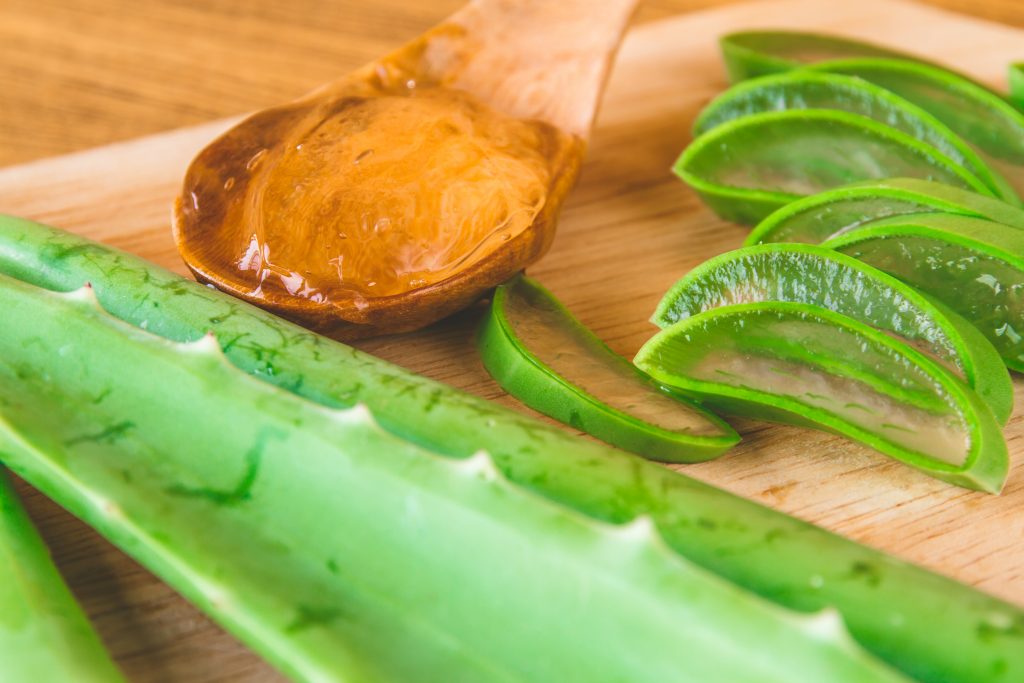 A wooden spoon full of aloe vera gel alongside pieces of the aloe vera plant against a wooden surface.