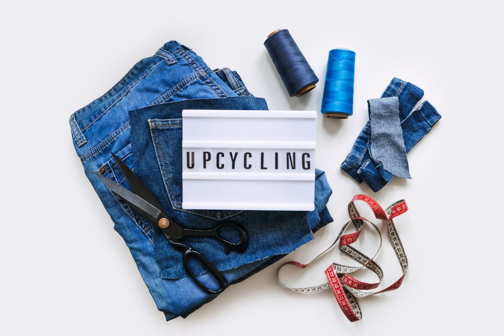 A pile of old jeans, spools of threads, measuring tape, scissors, and a plastic sign saying upcycling against a white background.