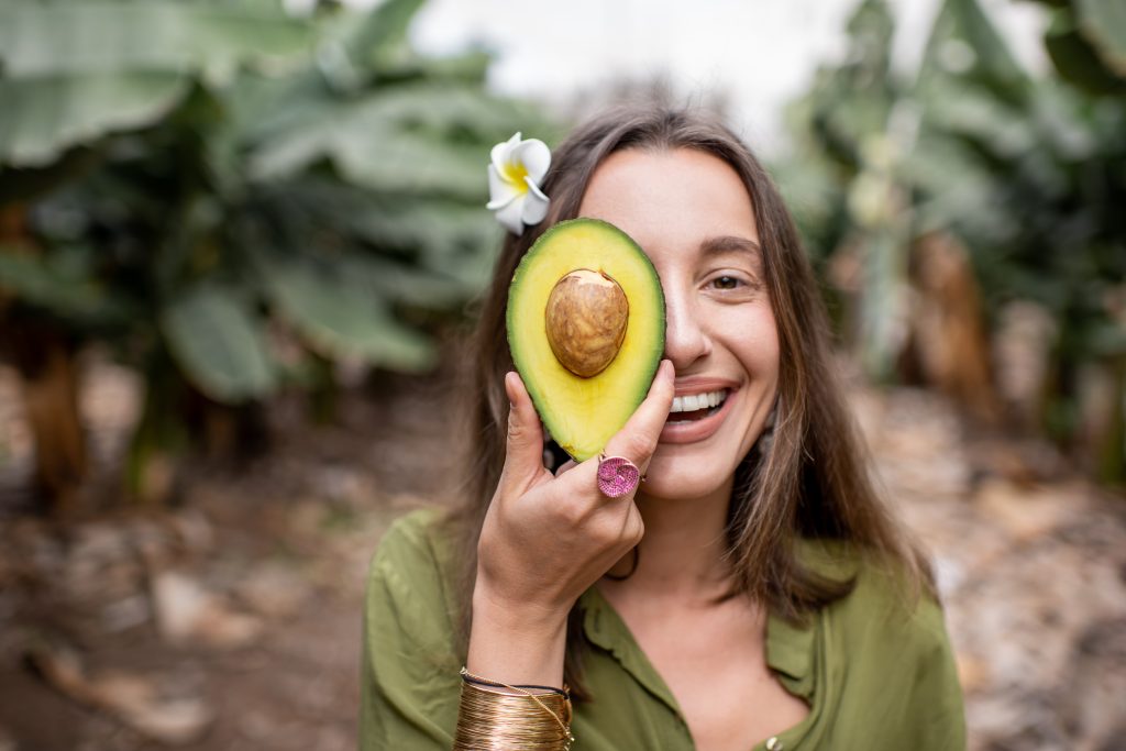 A smiling woman with clear, healthy skin standing outdoors and holding half of a cut avocado in front of her face.