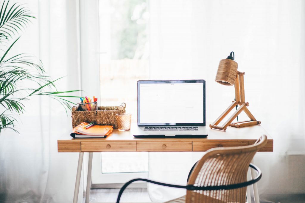 A home office desk near a window, with a laptop, office supplies basket, and lamp on the table. 