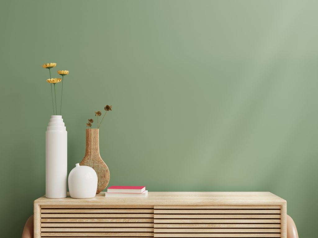 A green accent wall with a wooden cabinet as part of home decor. Flower vases and books are visible on top of the cabinet. 