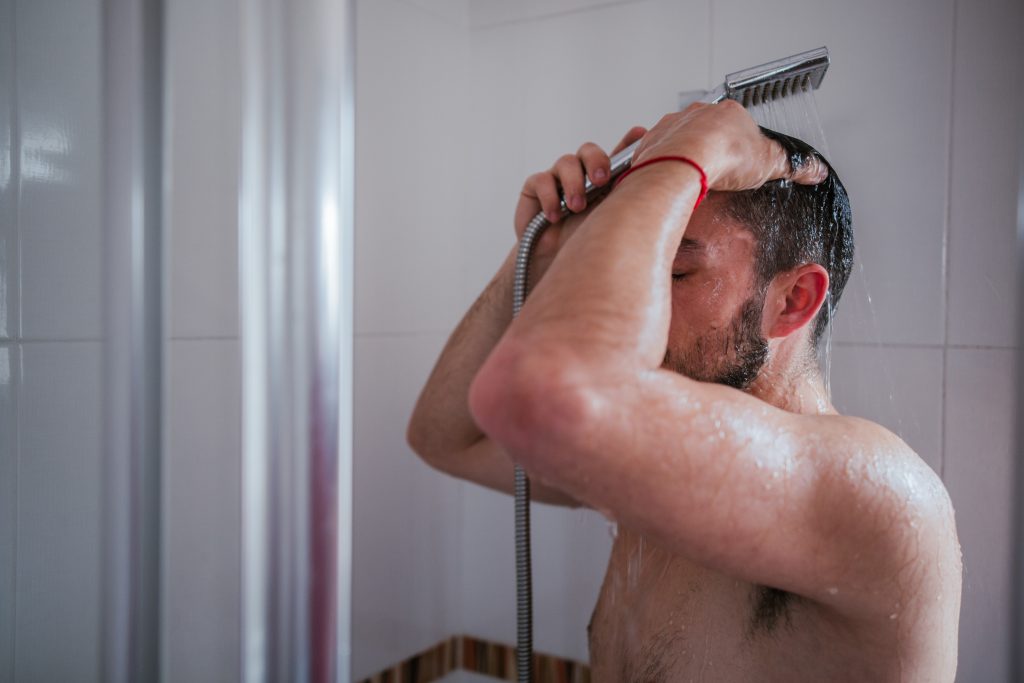 A man using a shower head to shower with hard water in a white bathroom. Showering with hard water can cause skin issues. 