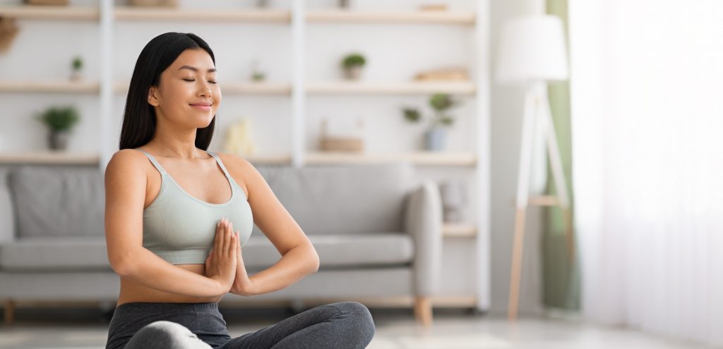 An Asian woman sits on a yoga mat and practices yoga at home.