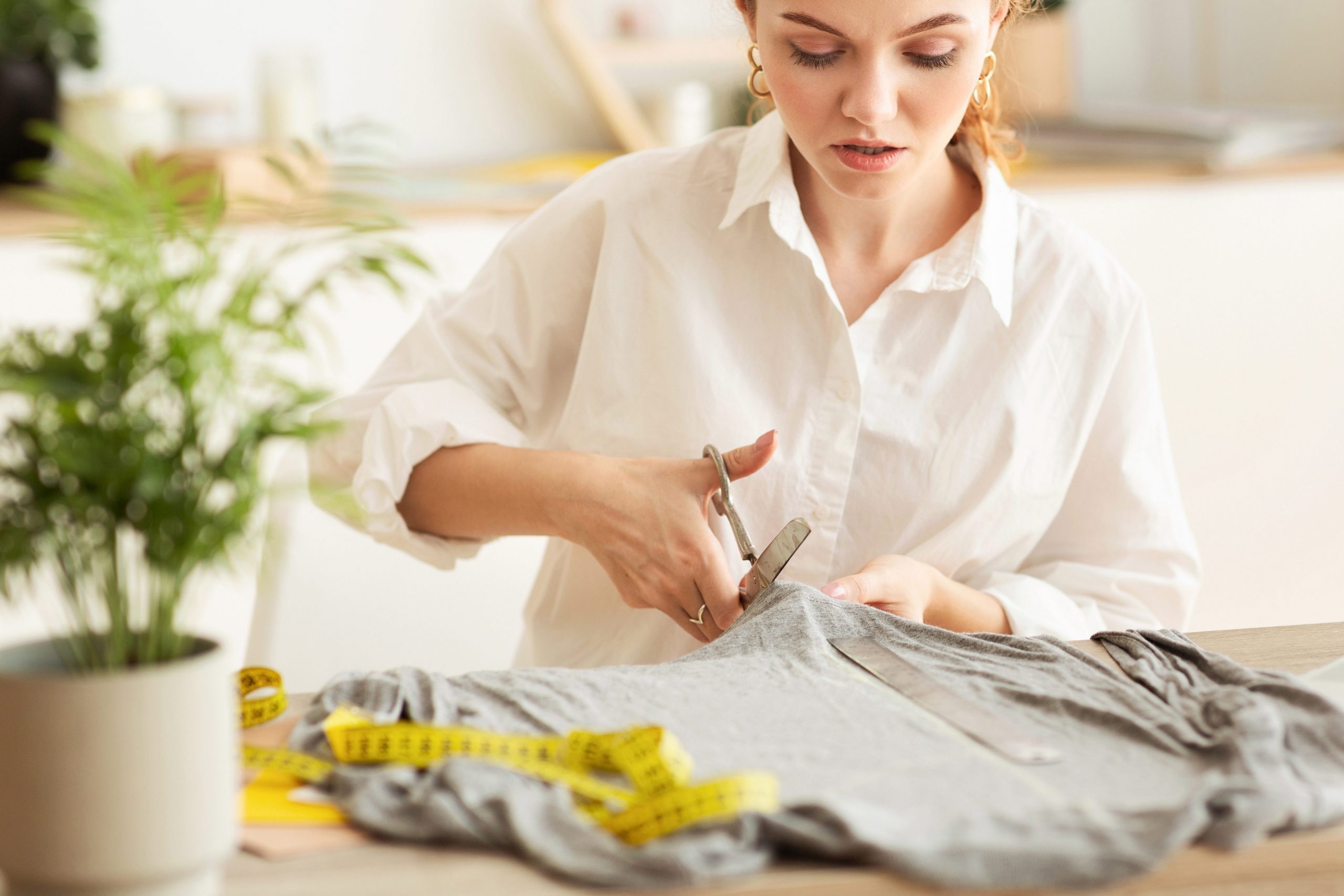 A woman in a white shirt sits at a desk and cuts an old t-shirt with scissors to repurpose and turn it into a cleaning rag. 