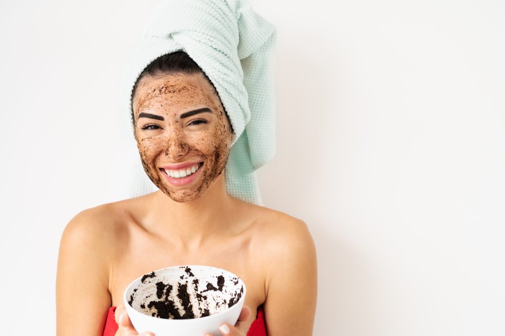 A woman with a hair towel holding a bowl of coffee grinds and showing her smiling face with coffee grinds on it. 