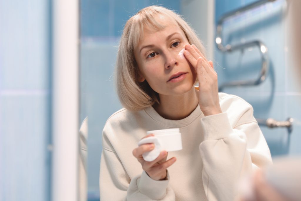 A woman with white hair applies moisturizer to her sensitive skin while looking into a bathroom mirror.