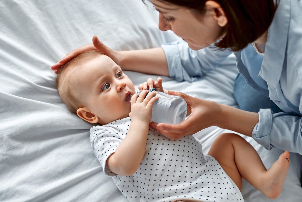 A mother and baby lying on a bed. Baby is drinking from a baby bottle.