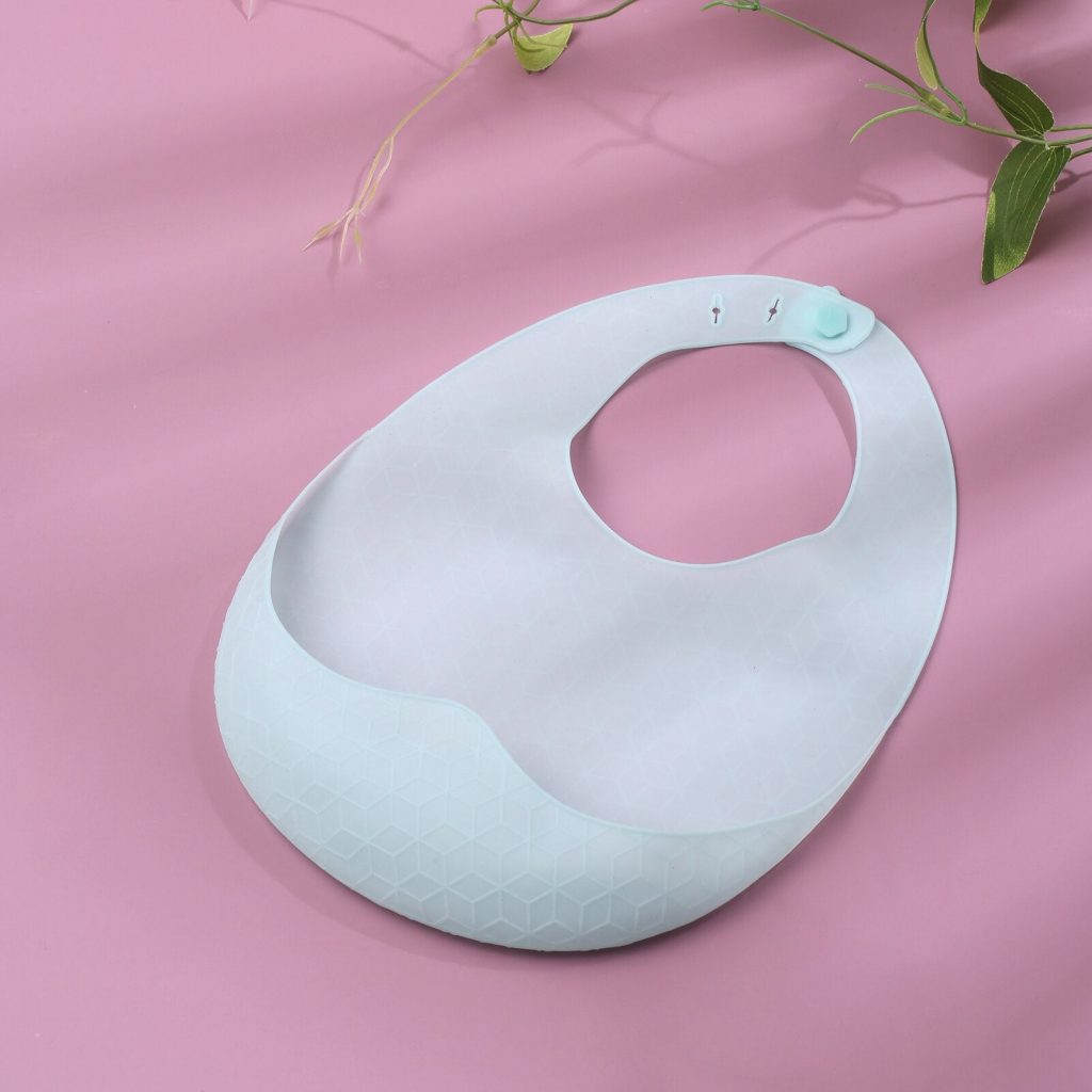 SuperTots Silicone Baby Bib in blue color against a pink background. Leaves visible in the top right corner.