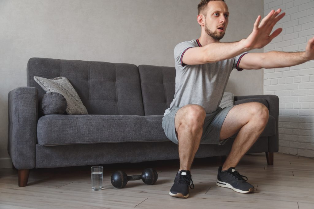 A young man does squats at home in front of his sofa. Glass of water and dumbbell on the floor. Concept of staying active indoors.