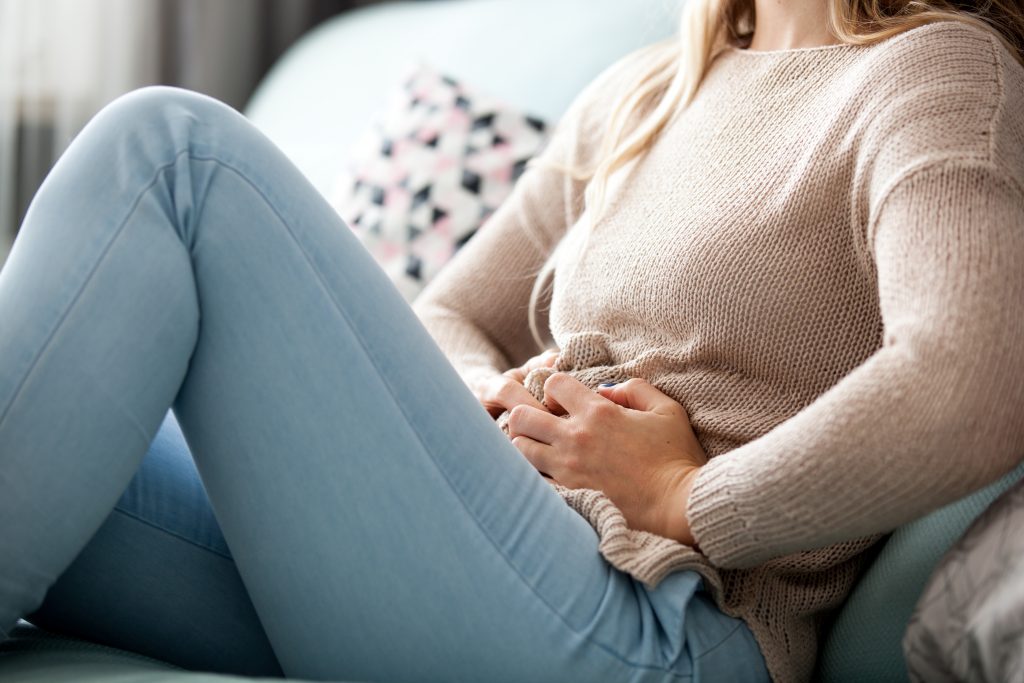 A young woman holding her stomach due to bloating. She is sitting on a safe, her face is not visible. Concept of bloating, bloating tips, how to reduce bloating.